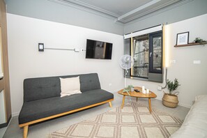 A modern living room with a grey sofa, a small wooden table, a floor lamp, and a large window with a view of the outside. - Bright Studio · Selected Design (H11) (Providencia)