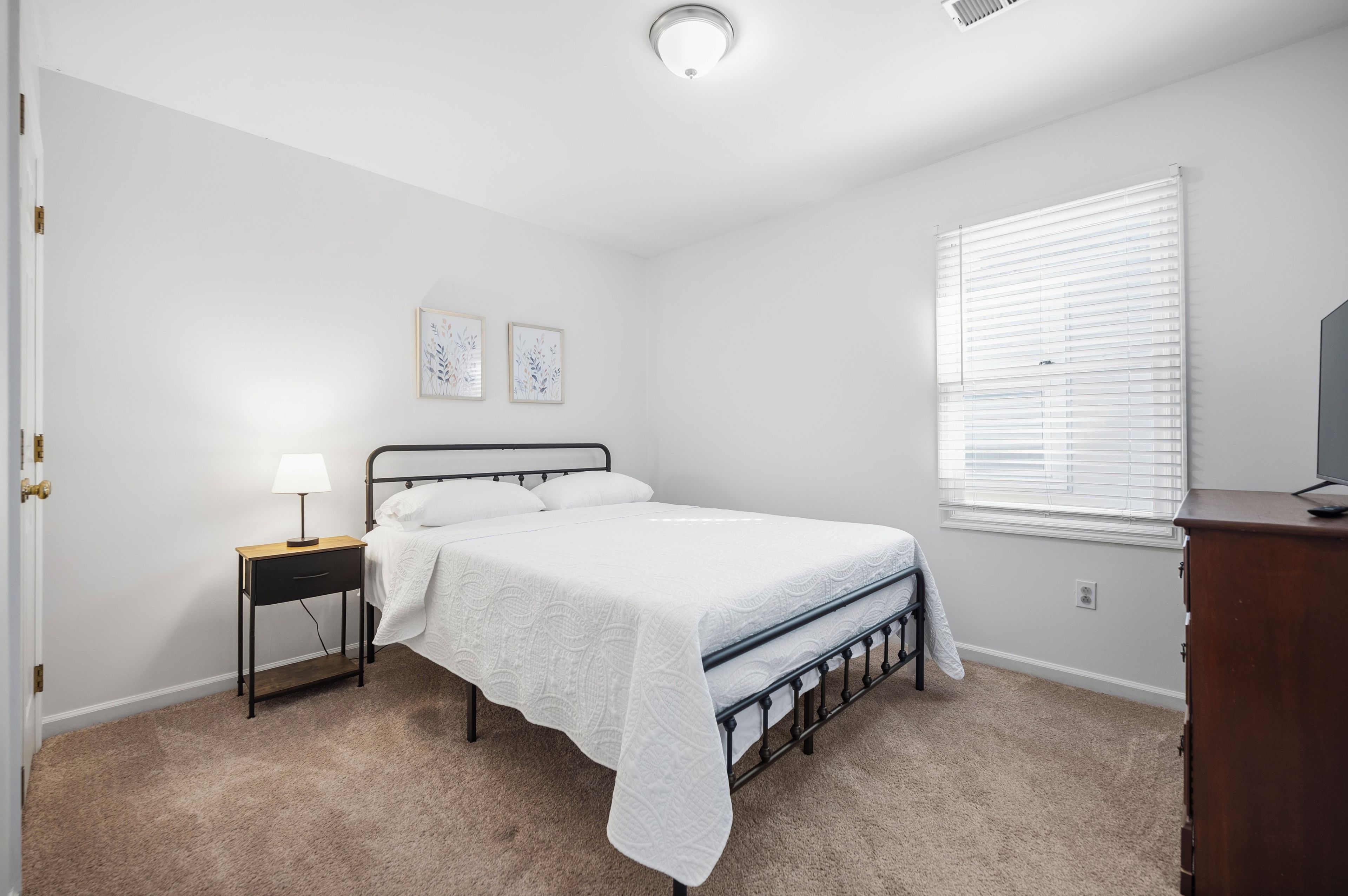 Ein Schlafzimmer mit Bett, Nachttisch, Lampe, zwei Wandbildern, einem Fenster mit Jalousien und einer Kommode.