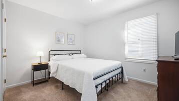 Ein Schlafzimmer mit Bett, Nachttisch, Lampe, zwei Wandbildern, einem Fenster mit Jalousien und einer Kommode.