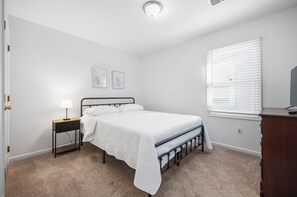 Un dormitorio con cama, mesita de noche, lámpara, dos cuadros en la pared, una ventana con persianas y un aparador.