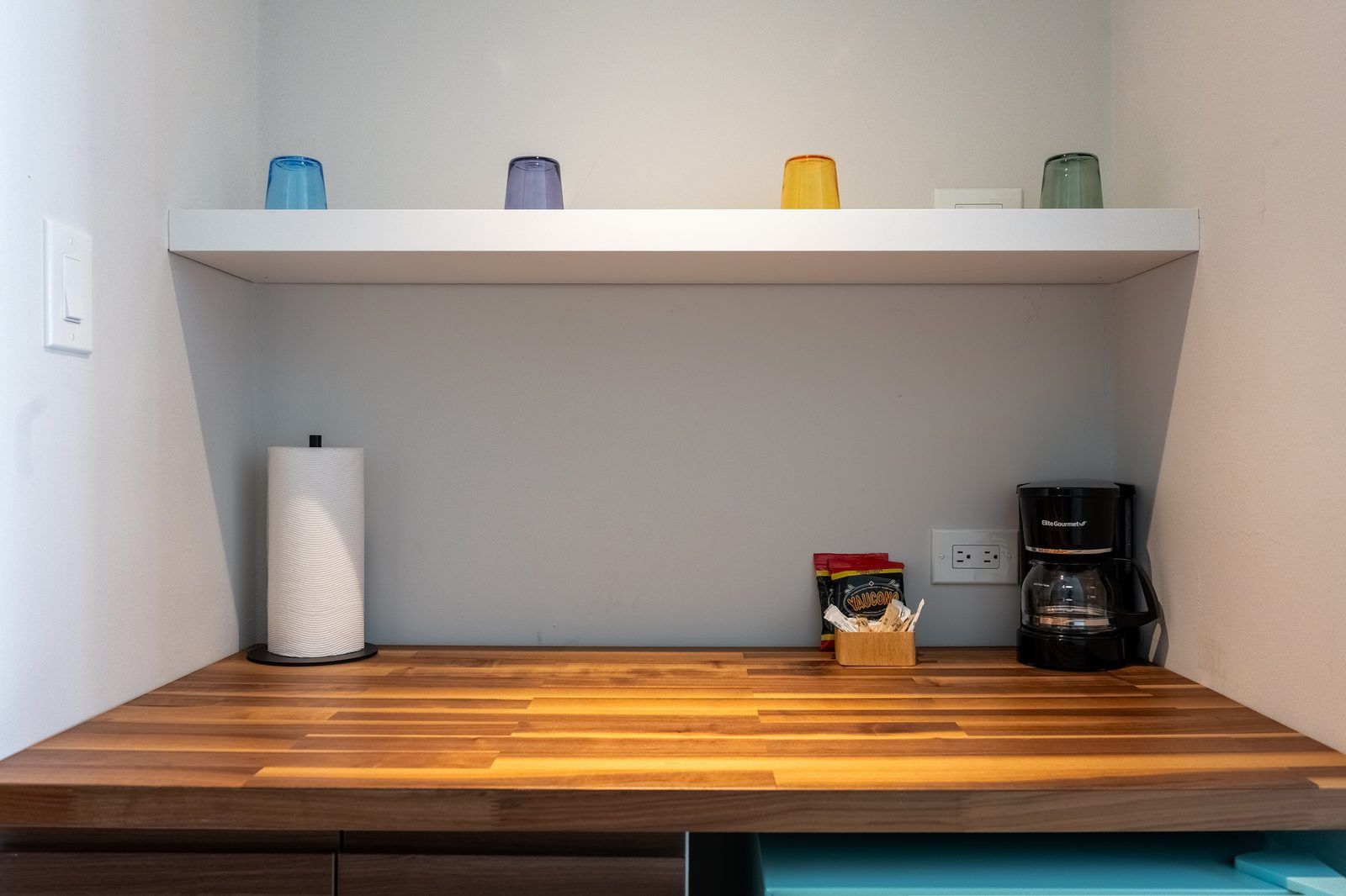 Un coin cuisine avec un plan de travail en bois, une étagère blanche ornée de verres colorés, une machine à café et un sac de collations.