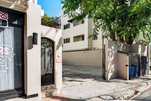 A residential street with a closed shop on the left, a metal gate with a no dogs sign, and a tree with dense foliage. - Central Sosua 1BR , Balcony , Hard Rock Perks (Sosúa)