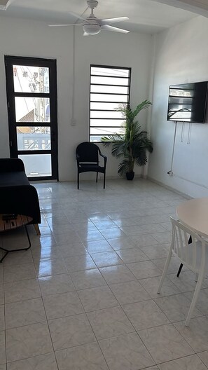 Una habitación amplia con ventilador de techo, un gran ventanal con persianas, una planta en maceta, un televisor de pantalla plana montado en la pared y una mesa de comedor con sillas.