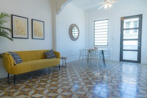 Une pièce avec un canapé jaune, une table à manger avec des chaises, un miroir et une porte avec une fenêtre.