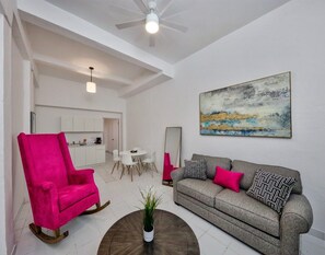 A living room with a bright ceiling fan, a sofa with patterned cushions, a vibrant pink chair, and a painting on the wall. - Chic Apt Near Old San Juan and Beach - Casa Pelayo 1 (San Juan)