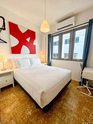 Een hotelkamer met een groot bed, twee nachtkastjes met lampen, een bureau met stoel, een raam met gordijnen en een rode abstracte schildering aan de muur.