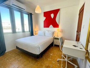A hotel room with a bed, bedside tables, a desk, and a large window.