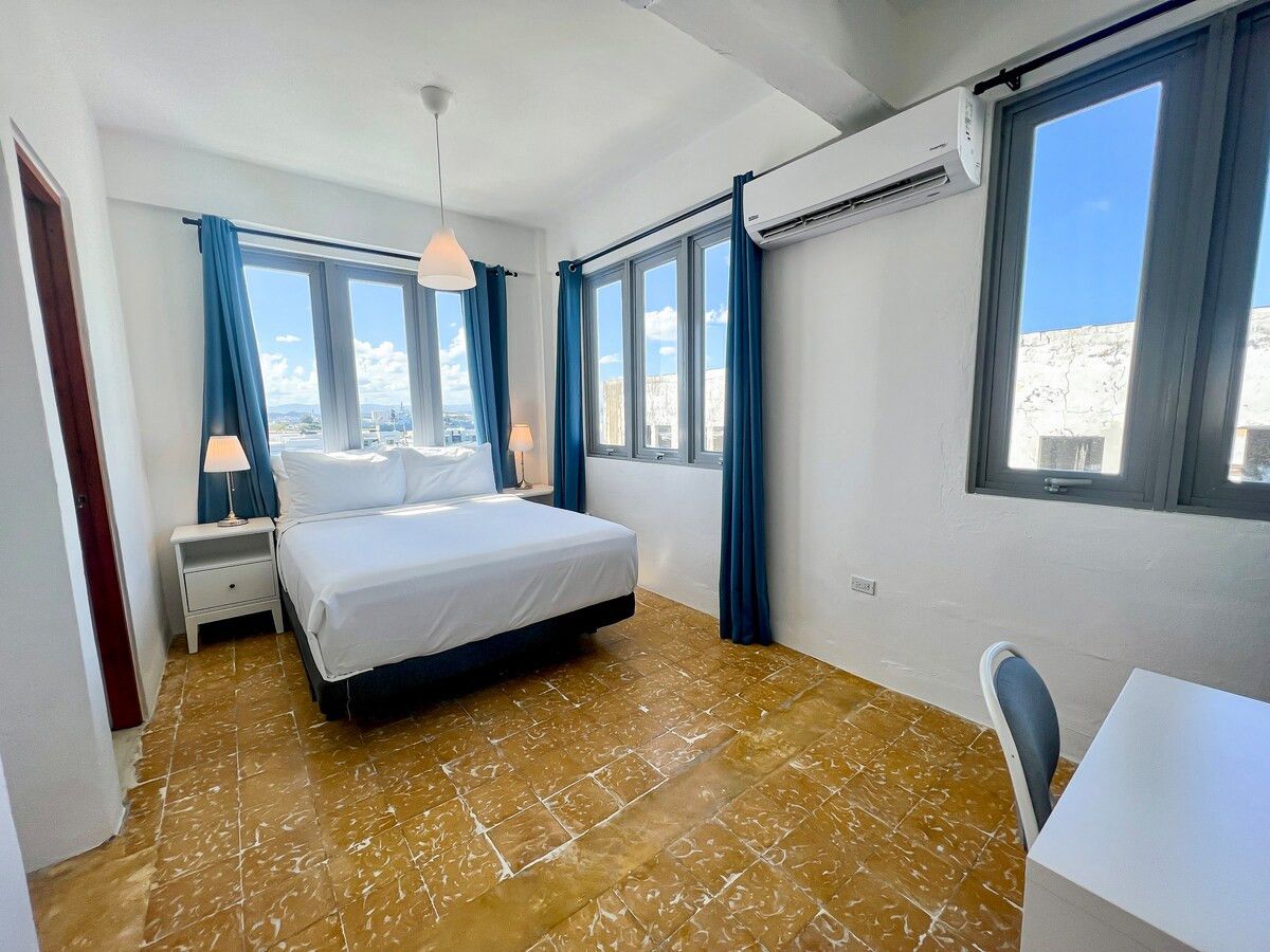 A hotel room with a bed, a desk, a chair, and large windows.