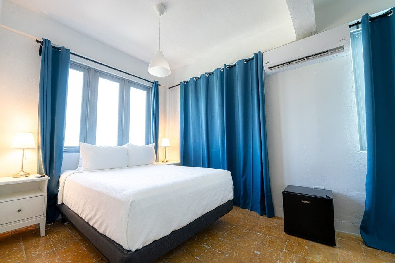 Une chambre d’hôtel comprenant un lit, deux tables de chevet, une lampe, une fenêtre avec des rideaux, un appareil de climatisation et un petit haut-parleur noir.