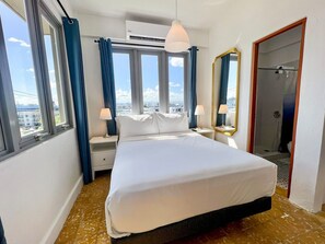 Een hotelkamer met een groot bed, twee nachtkastjes, een spiegel en uitzicht op de stad door de ramen.