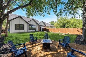 A backyard with a fire pit, four Adirondack chairs, a wooden deck, and a house with a sloped roof and large windows. - Spacious Dallas Retreat - Sleeps 10 - Near Downtown & Trinity Groves (Dallas)