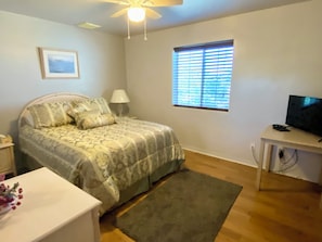 Ein Schlafzimmer mit einem Bett, einem Schreibtisch mit Fernseher, einem Fenster mit Jalousien, einem Deckenventilator und einem Nachttisch mit Lampe.