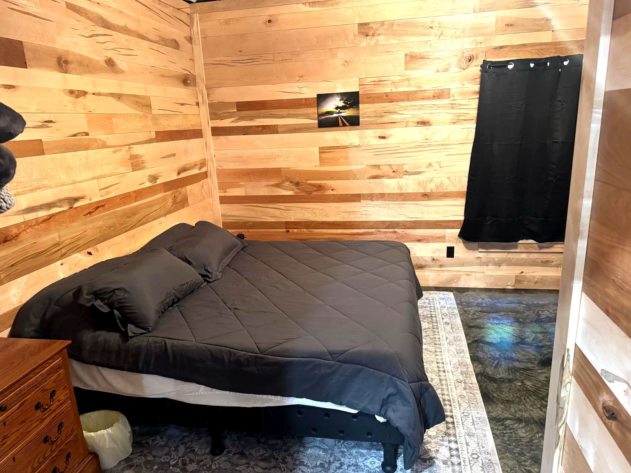 A wooden room with a bed, a small bedside table, a picture on the wall, and a black curtain.