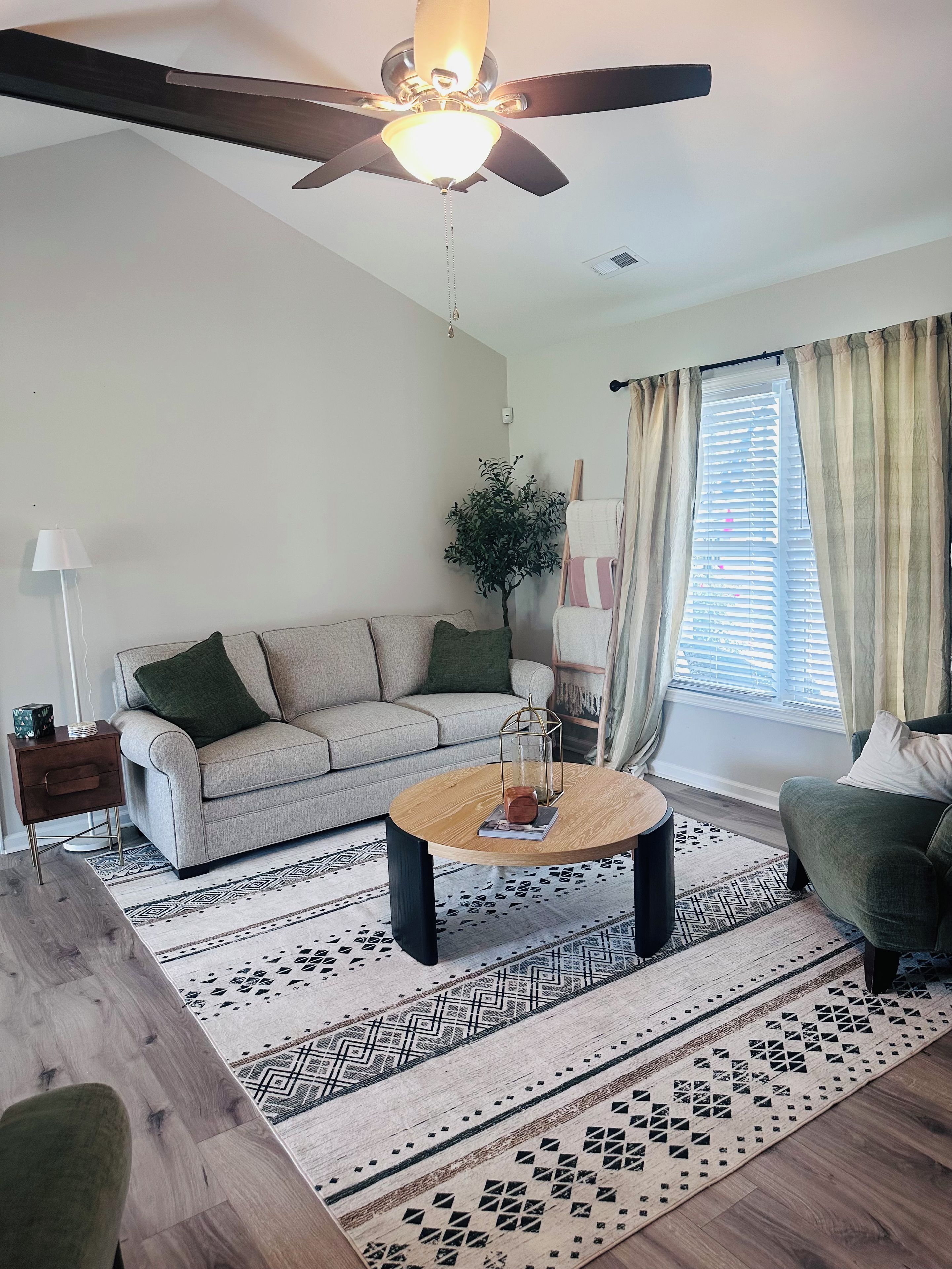 A living room with a ceiling fan, a sofa, a coffee table, a lamp, and a window with curtains.