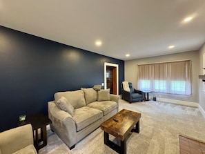 A living room with a dark blue wall, a beige sofa, a black leather chair, a wooden coffee table, and a window with curtains. - NEW! Stylish Stay in the Heart of Palmyra! (Palmyra)