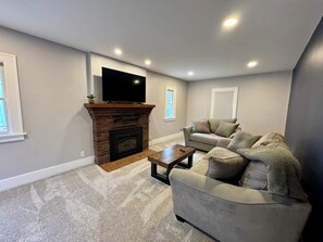 A living room with a fireplace, a flat-screen TV mounted above it, a grey sofa, and a coffee table. - NEW! Stylish Stay in the Heart of Palmyra! (Palmyra)