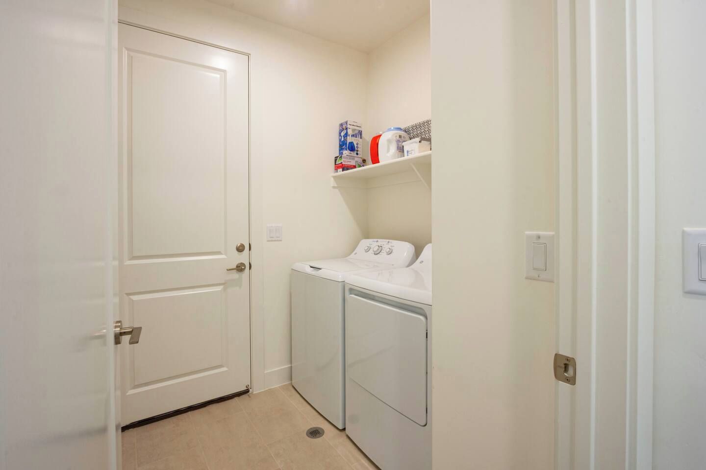A laundry room with a washing machine and dryer, a door, and shelves with laundry supplies.