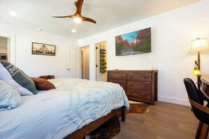 Une chambre à coucher avec un lit, une commode en bois, un ventilateur de plafond, un tableau et une lampe.