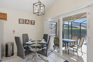 A dining area with a glass table and chairs, a sign with the word 'EAT' on the wall, and a view of an outdoor pool through the glass door. - Amazing Retreat at Lake Berkley (Kissimmee)