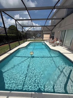 A swimming pool with a clear blue water surface, surrounded by a covered area with lounge chairs and a view of a house in the background. - Amazing Retreat at Lake Berkley (Kissimmee)