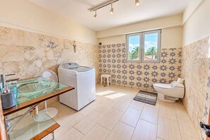 Un baño con lavamanos con recinto de vidrio, lavadora, inodoro y ventana con vista a palmeras.