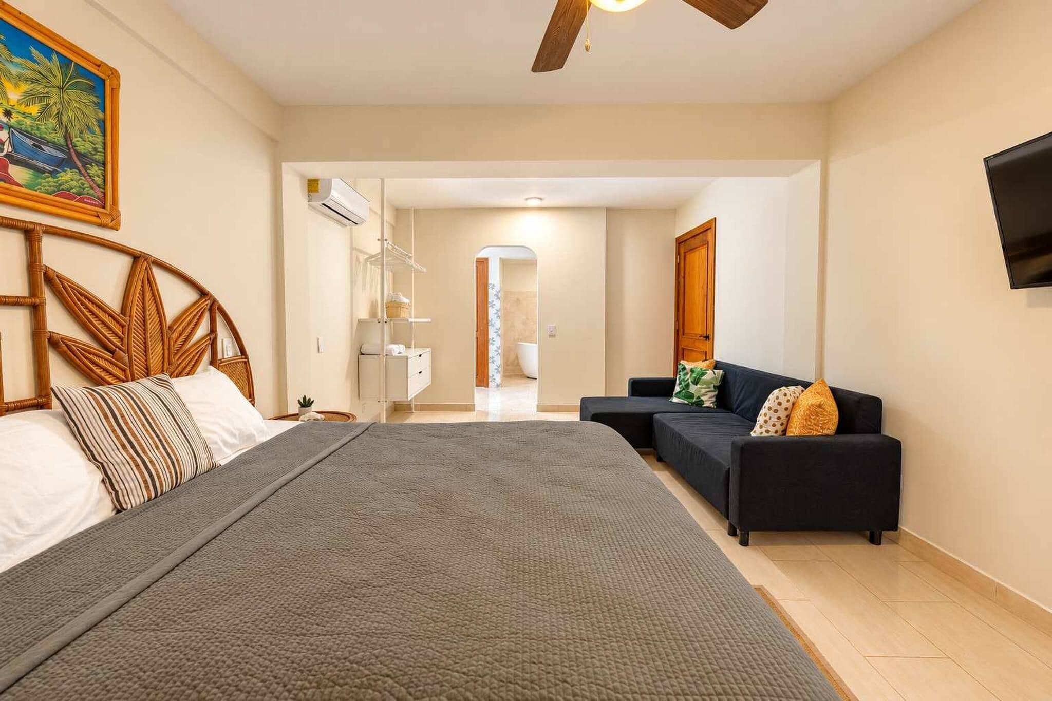 Un dormitorio con cama, sofá, televisión y baño visible a través de una puerta abierta.