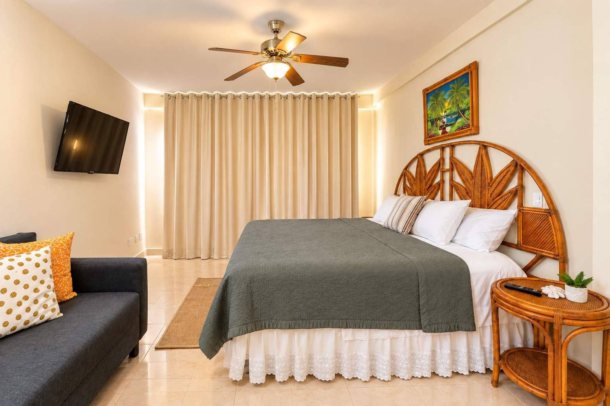 Un dormitorio con cama, televisor, ventilador de techo, un cuadro, sofá y una pequeña mesa.