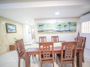 A dining area with a wooden table and chairs, a painting on the wall, and a potted plant. - Spacious 1BR APT in Sosua Strip! (Sosúa)