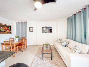 A living room with a sofa, coffee table, dining table, chairs, a TV, and a painting. - Secure and Central 2BR Apt (Sosúa)