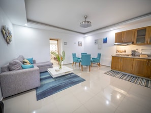Ein modernes Wohnzimmer mit einem grauen Sofa, einem Couchtisch und einem blauen Teppich. Es gibt einen Essbereich mit blauen Stühlen und eine Küche mit Holzausbau.
