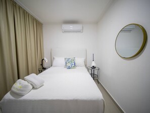 A hotel room with a bed, a bedside table with a lamp, a wall-mounted air conditioner, and a round mirror. - 2BR APT in Sosua Center Strip Zone (Sosúa)