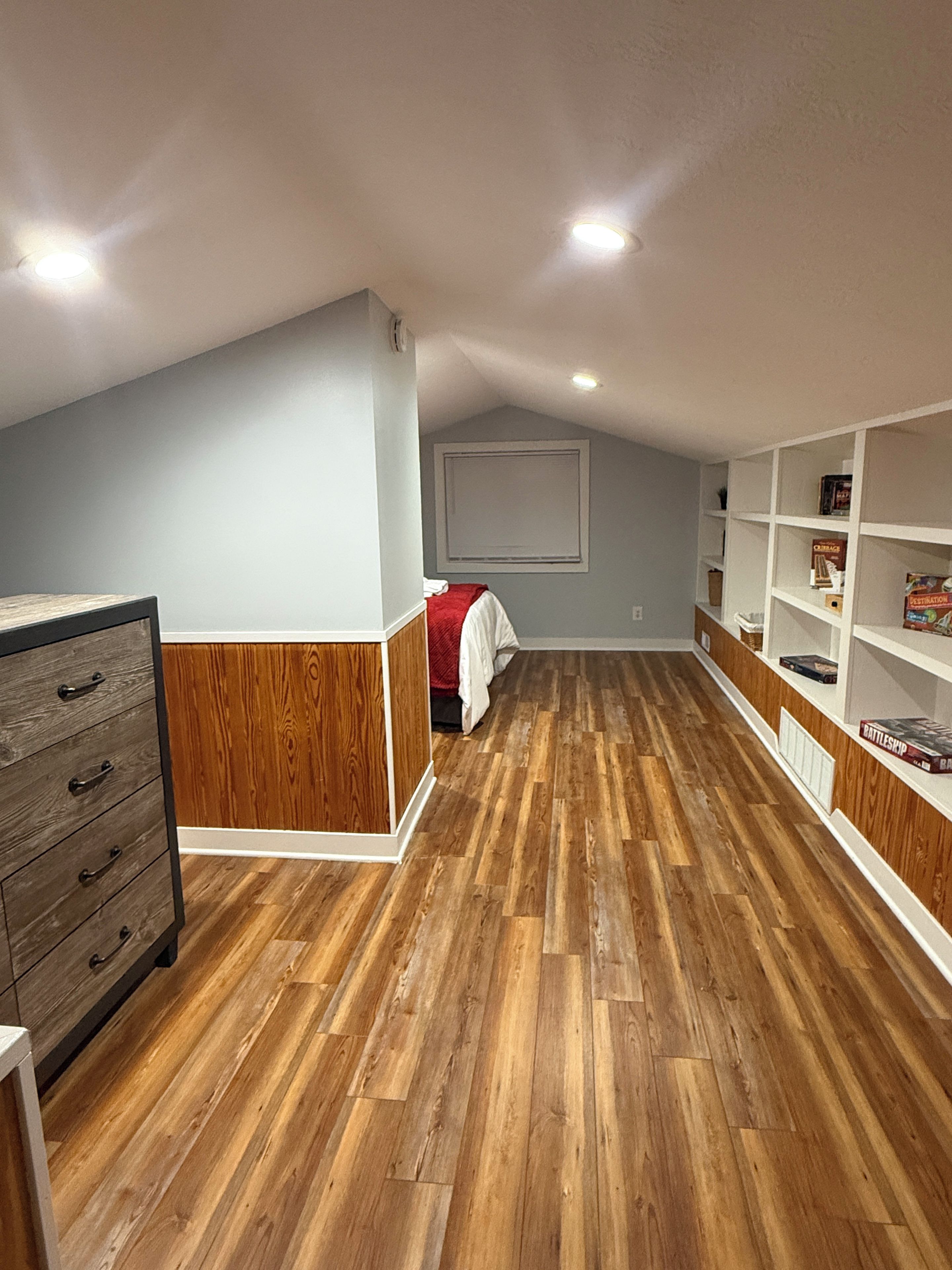 A room with wooden flooring, a dresser, a bed with a red blanket, and built-in shelving with books.
