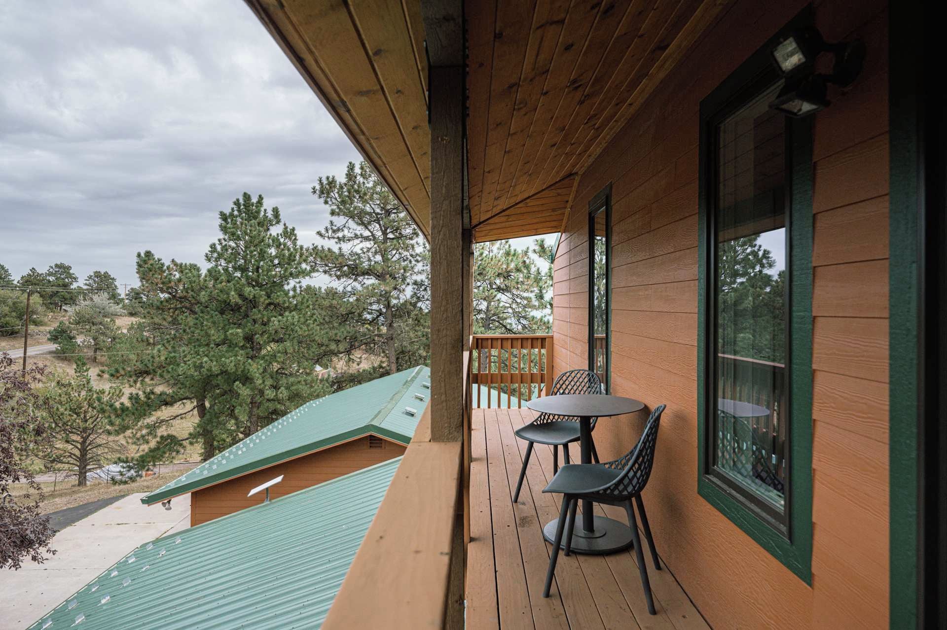 A wooden deck with a table and chairs, overlooking a green-roofed building and a forested area.
