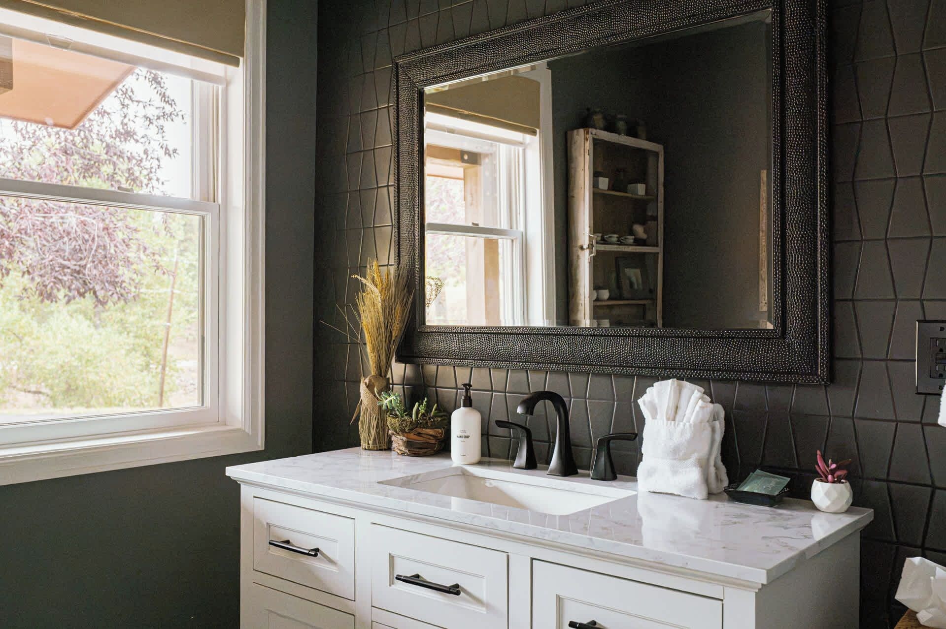 A bathroom with a white vanity, a black-framed mirror, and a window with a view of trees.