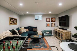 Uma sala de estar com sofá, mesa de pebolim, mesa de centro e televisão na parede.