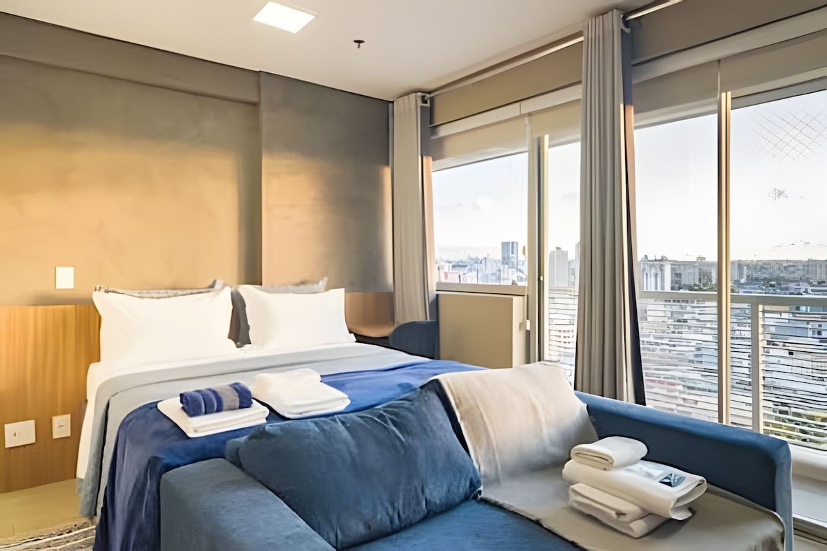 A modern hotel room with a large bed, a sofa, and a view of a cityscape through large windows.