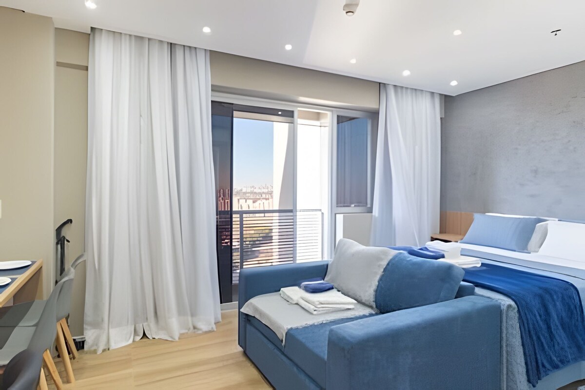 A modern bedroom with a bed, a desk, and a view of the city through a balcony.