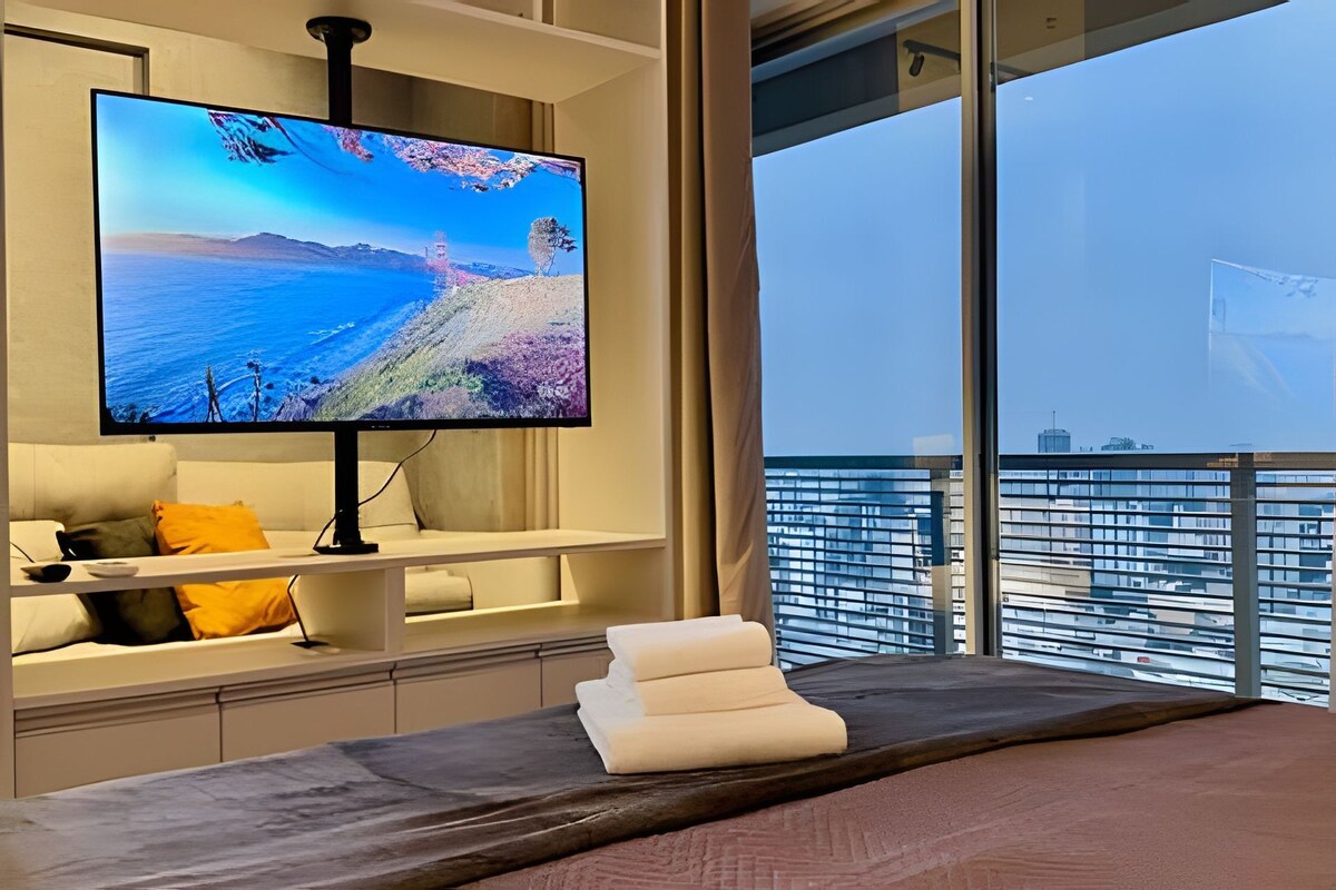 A hotel room with a large flat-screen TV on a stand, a bed with a dark headboard, a small table with towels, and a view of a cityscape through large windows.