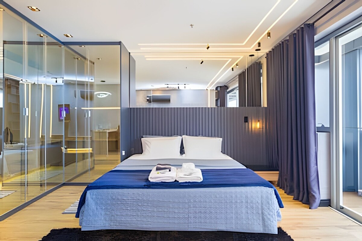 A modern hotel room with a large bed, a glass partition, and a view of the city.