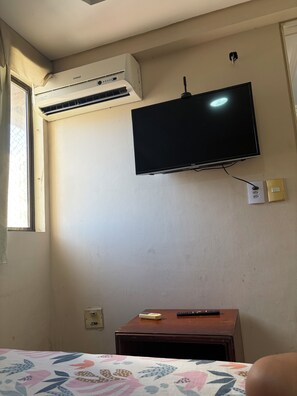 A room with a bed, a television mounted on the wall, an air conditioning unit, a small wooden table with a remote control, and a power outlet. - Apartment on the beachfront in Fortaleza (Fortaleza)