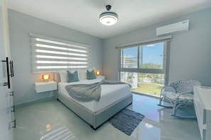 A modern bedroom with a bed, bedside tables, a chair, a window, and an air conditioning unit. - Exclusive Oceanfront 3 BR Apt in Sosua (Sosúa)