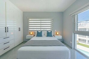 Une chambre à coucher avec un grand lit, deux tables de chevet avec des lampes, une fenêtre avec des stores et une vue sur un bâtiment à l’extérieur.
