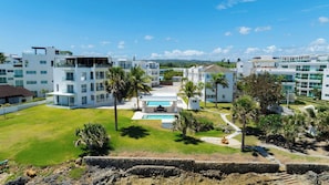 A modern hotel complex with multiple white buildings, a central swimming pool, and surrounding palm trees.