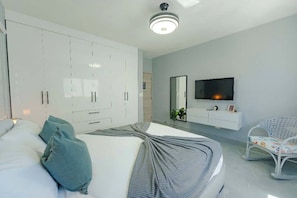 A modern bedroom with a bed, white wardrobe, TV, and a small seating area.