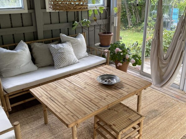 Un banco de madera con asiento de mimbre, una mesita con un cuenco, una planta en maceta y un banco acolchado con cojines.