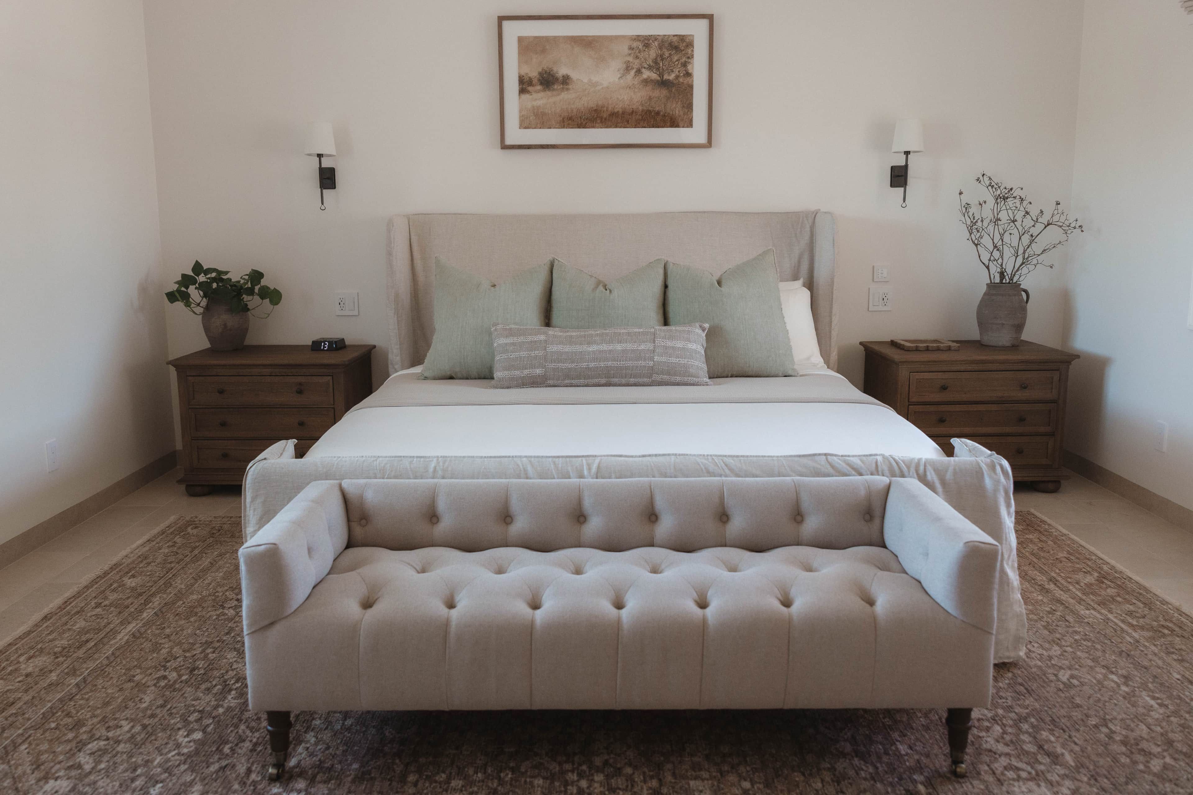 A bedroom with a large bed, two nightstands, a bedside table, a bench, and a wall-mounted lamp.