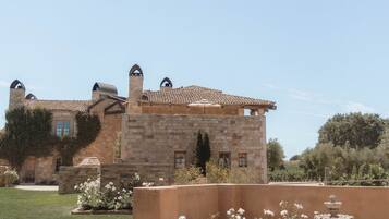 Edifici de pedra amb finestres d'arc i teulat de rajola, envoltat d'un jardí amb flors blanques i una font.