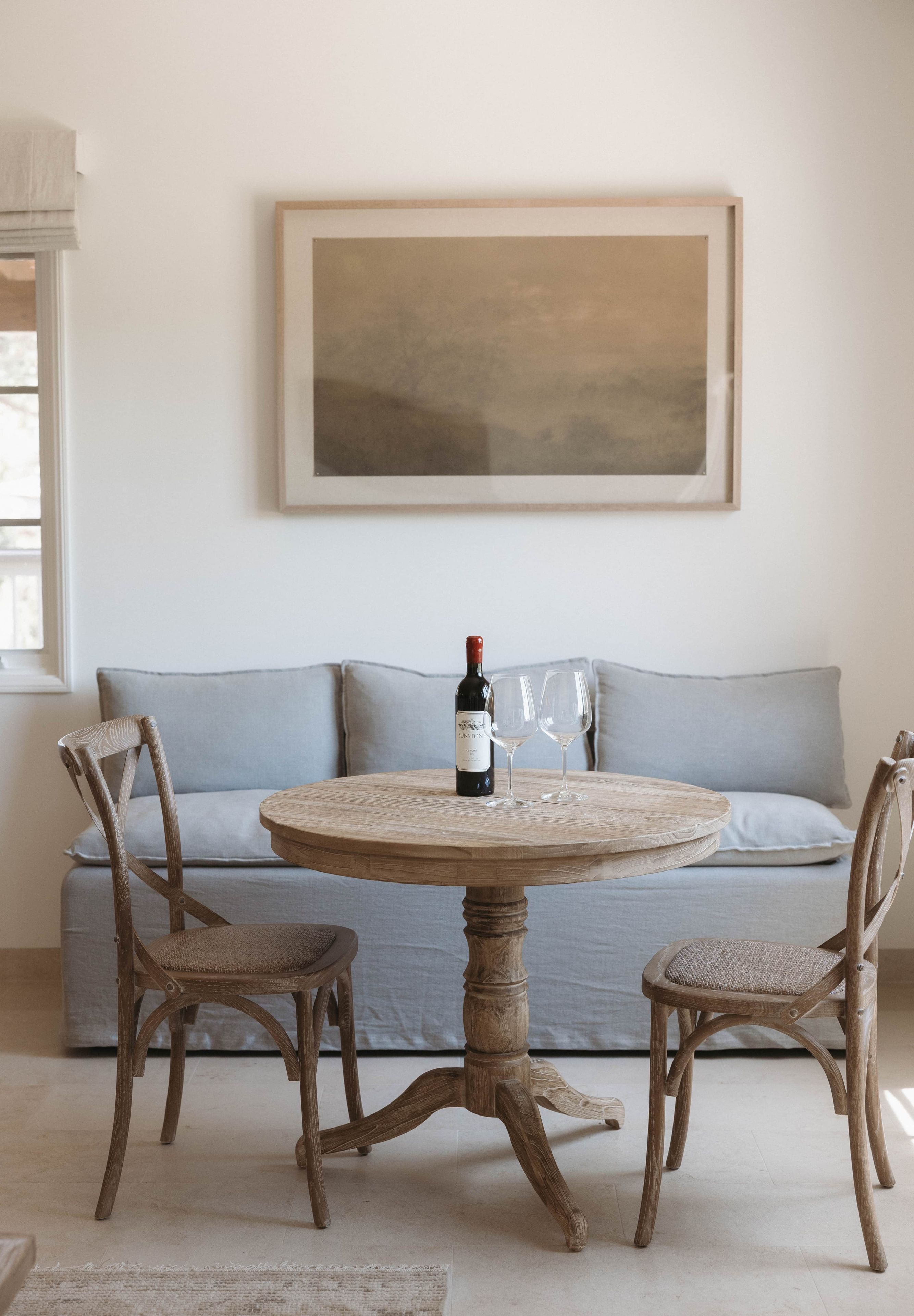 A round wooden table with a bottle of wine and two glasses, surrounded by wicker chairs, in a room with a light blue sofa and a large abstract painting on the wall.