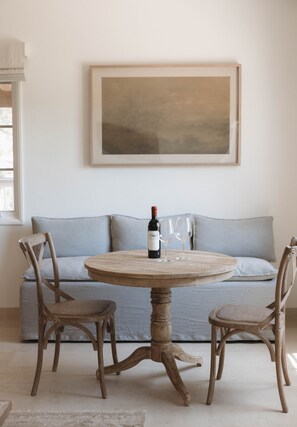 A round wooden table with a bottle of wine and two glasses, surrounded by wicker chairs, in a room with a light blue sofa and a large abstract painting on the wall. - Sunstone Cottage the Champagne Suite (Santa Barbara County)