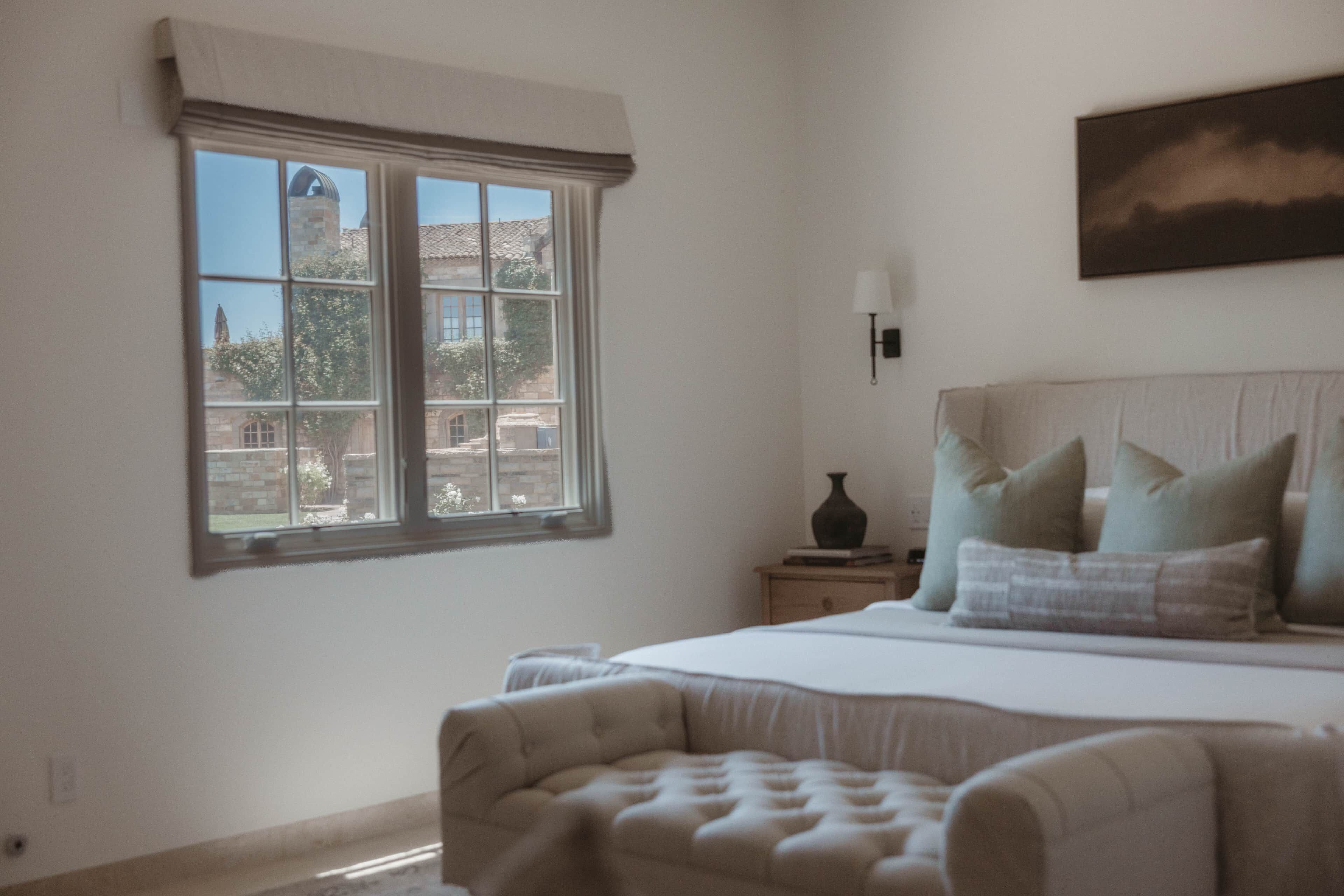 A bedroom with a large bed, a nightstand, a lamp, and a window with a view of buildings.